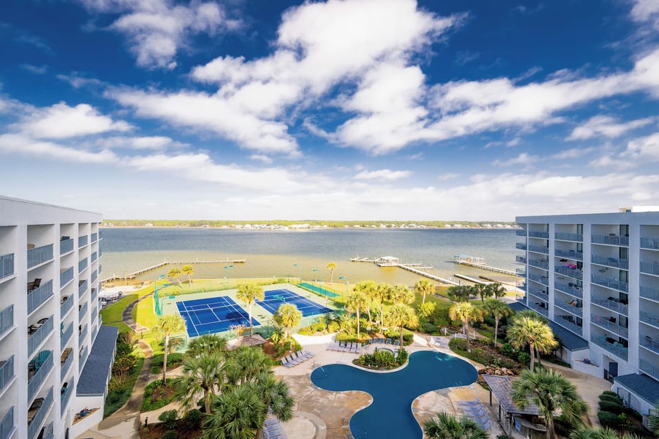 This stunning view showcases a serene waterfront with lush palm trees, a pool, and tennis courts, perfect for relaxation