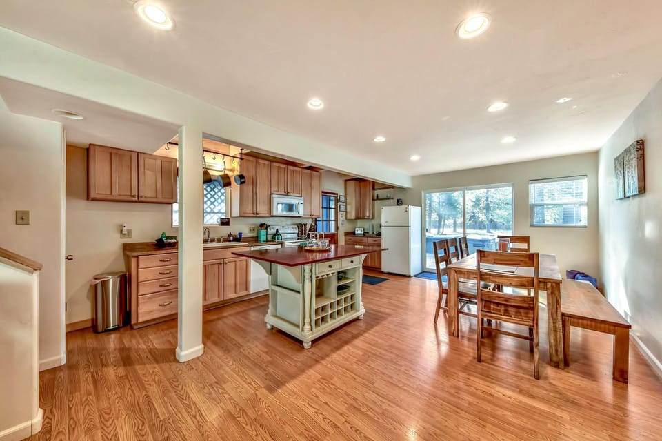 Dining area with wooden table, bench seating, and large windows next to an open kitchen with center island.