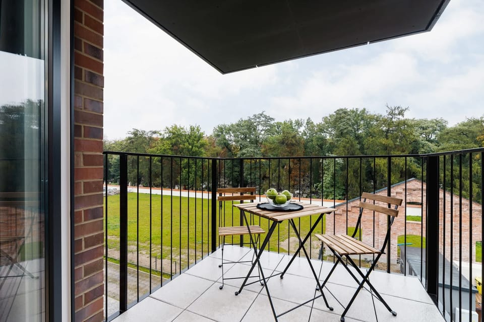 A small balcony with a simple black table and two chairs, overlooking a green outdoor area.