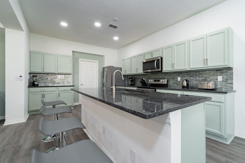 Modern kitchen with mint green cabinetry, large granite island, breakfast bar seating, stainless steel appliances, tile backsplash, and recessed ceiling lighting.