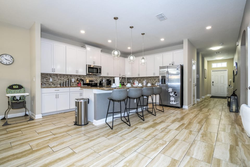 Open kitchen with white cabinets, mosaic tile backsplash, granite countertop island, and four gray bar stools.
