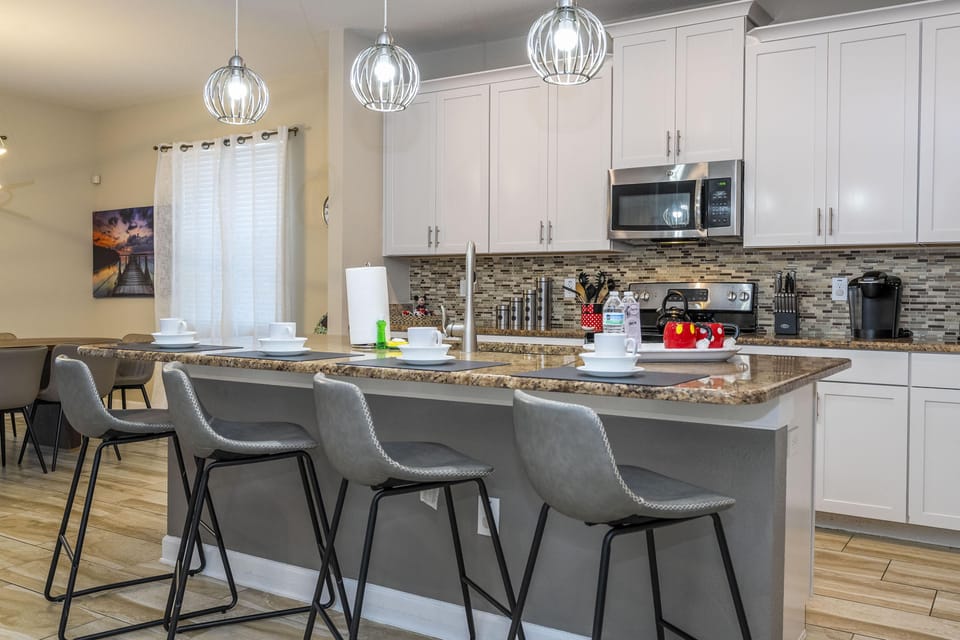 Granite countertop island with four gray barstools beneath three pendant lights in the kitchen.