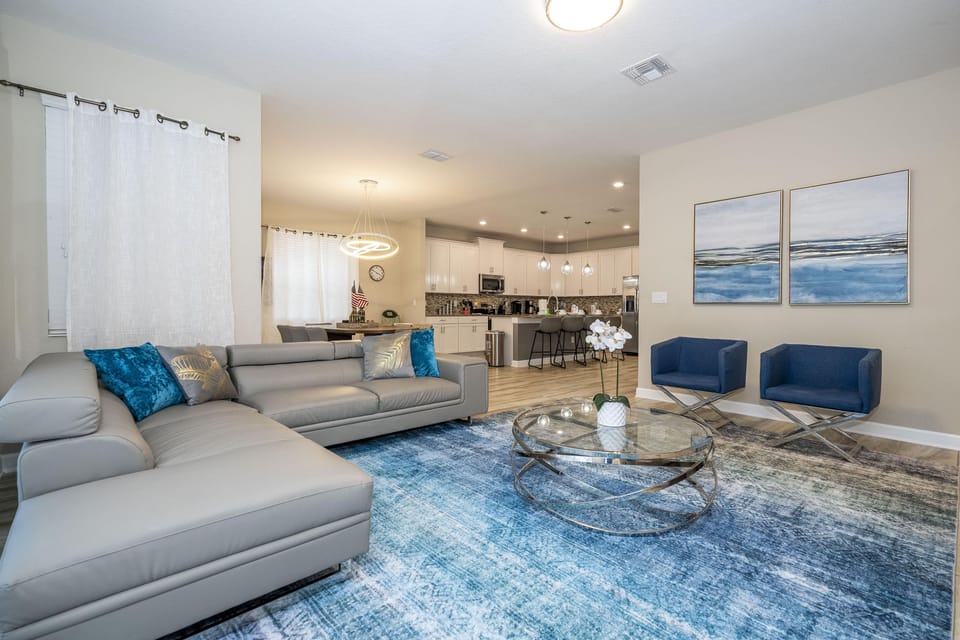 Living room with gray leather sectional, blue accent chairs, glass coffee table, and abstract ocean-themed wall art.