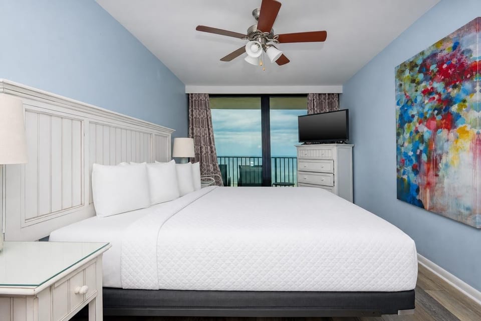 This bedroom features a king-sized bed, a colorful abstract painting, and a TV, with a stunning beachfront view