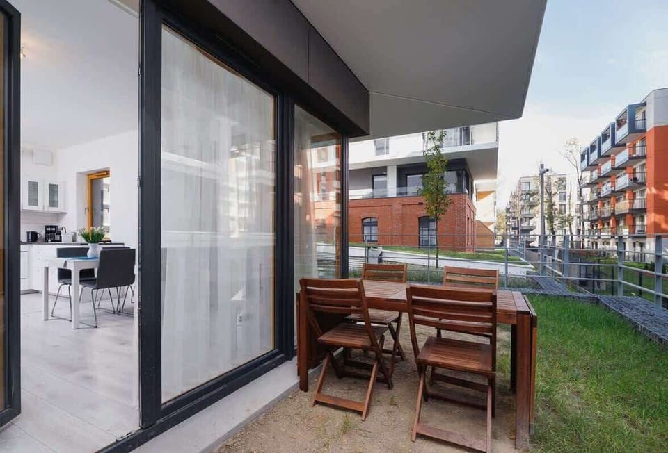 A small terrace with a wooden table and chairs set, overlooking a garden area. It has sliding glass doors leading to the interior of the apartment.