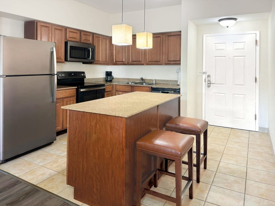 Kitchen with wooden cabinetry, marble-finish countertops, stainless fridge and oven, and island bar stools