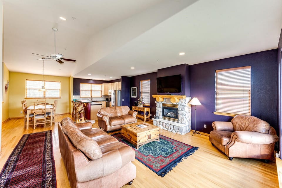 A spacious living room with wooden flooring, featuring leather sofas, a stone fireplace, a wooden coffee table, and a wall-mounted TV. There's a dining area and kitchen visible in the background.