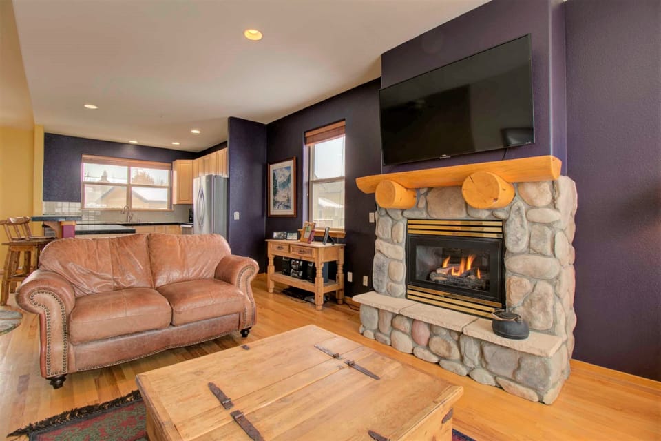 A cozy living room with a stone fireplace, leather armchair, and wooden coffee table. Mounted TV above the fireplace, kitchen and dining bar visible in the background. Walls painted dark purple.