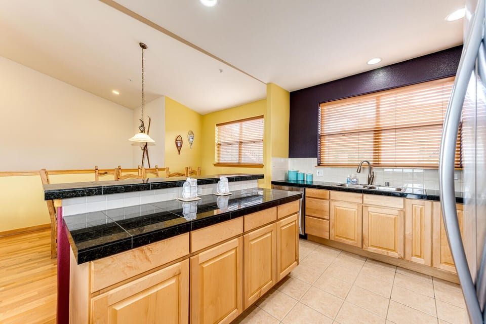 A modern kitchen with light wood cabinets, black countertops, a double sink, and stainless steel appliances. It features large windows with blinds and tiled flooring. The dining area is adjacent.