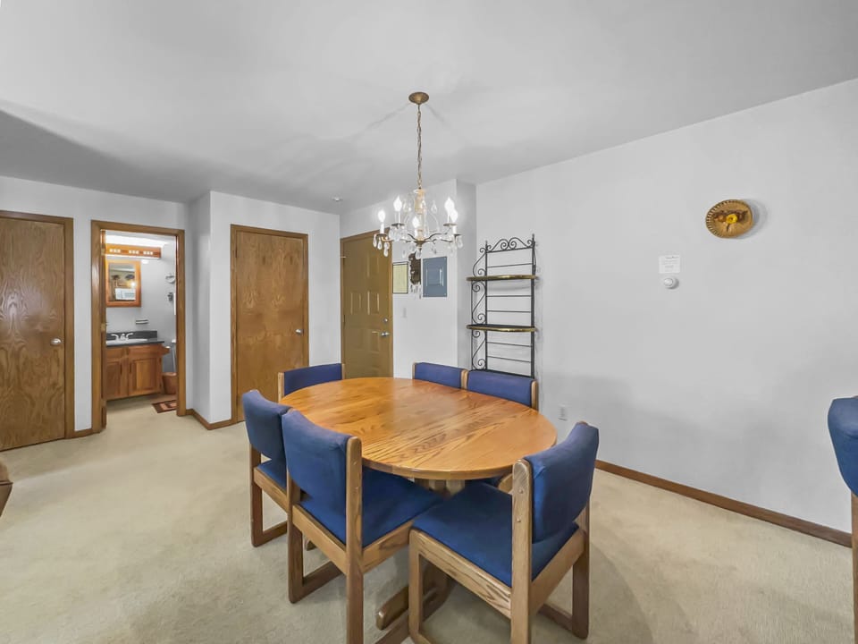 A dining area with a wooden table and six blue chairs under a chandelier. The room has a carpeted floor, three wooden doors, a metal shelf, and a small wall-mounted decoration.