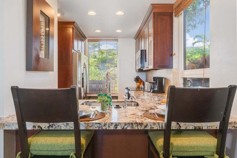 A modern kitchen with granite countertops, stainless steel appliances, and a breakfast bar set for two, filled with natural light and tropical garden views.