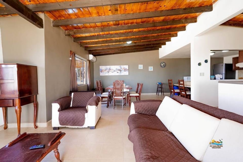 The living room features comfortable sofas, a classic coffee table, a TV hidden in a wooden entertainment center, and exposed wood beam ceilings with crisp white tile floors — an authentic Mexican beachfront getaway