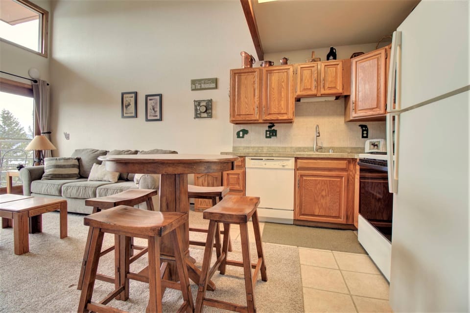 A cozy living area with a wooden dining set, light-colored sofa, and a compact kitchen featuring wooden cabinets, a white refrigerator, and a dishwasher. Decor includes framed pictures and a wall sign.