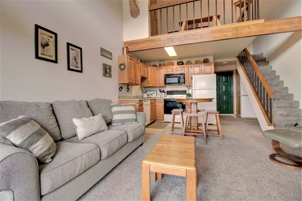 A cozy living room features a grey sofa, coffee table, kitchen with wooden cabinets, black appliances, a dining area, and a staircase leading to an upper level.