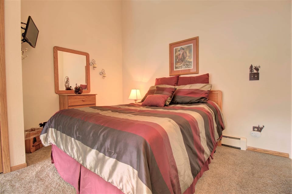 A bedroom with a neatly made bed featuring striped bedding, a wooden nightstand with a small lamp, a dresser with a mirror, and a wall-mounted TV. The room has beige walls and carpeted flooring.