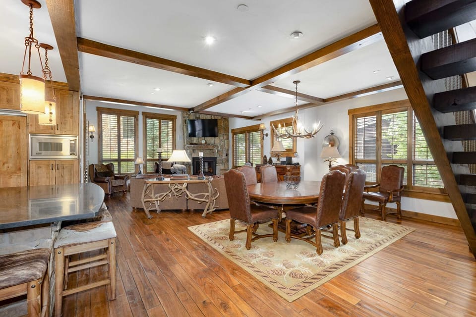 Open-concept living and dining area with wooden floors, a large dining table, a stone fireplace, and windows allowing natural light in. A staircase is visible on the right.