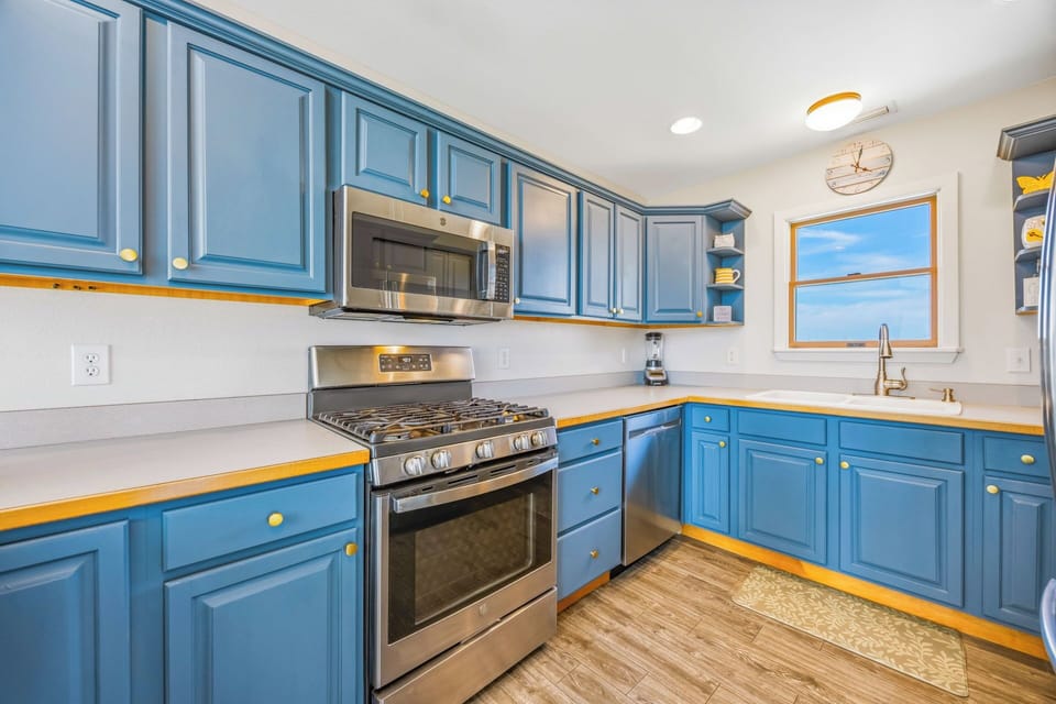 Your fully equipped kitchen features cheerful blue cabinetry, stainless appliances, and plenty of counter space—perfect for preparing home-cooked meals while enjoying ocean views through the window.