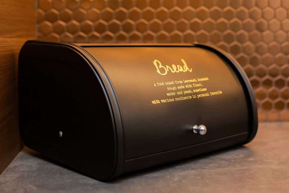 A close-up of a stylish, black bread box labeled "Bread," placed on a wooden kitchen counter.
