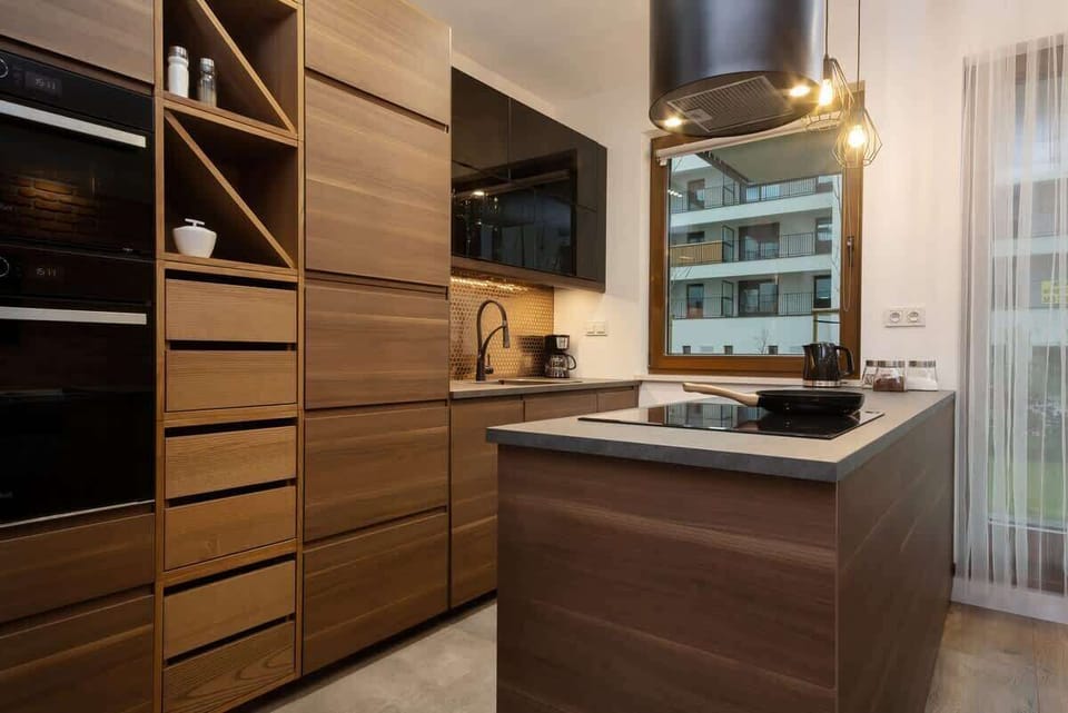 A modern kitchenette with dark wooden cabinets, built-in oven, and a black sink. The backsplash features a stylish black mosaic, adding a touch of elegance.