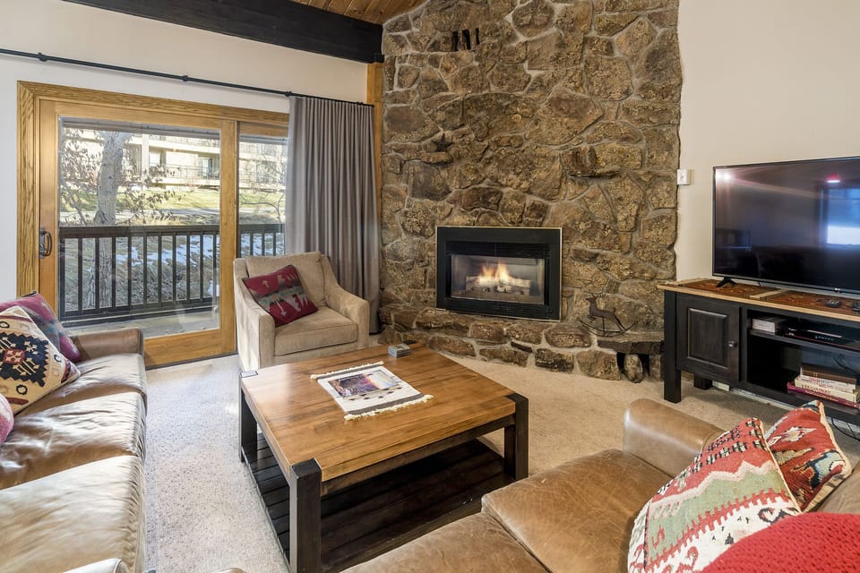 A cozy living room with a stone fireplace, leather sofas, a wooden coffee table, an armchair, a TV on a stand, and a sliding glass door leading to a balcony with a view of trees.