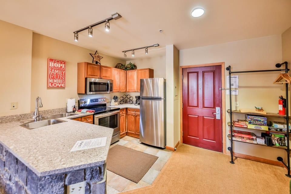 A cozy kitchen with wooden cabinets, stainless steel appliances, and a granite countertop. A red front door is on the right, and a bookshelf with games is against the wall next to it.