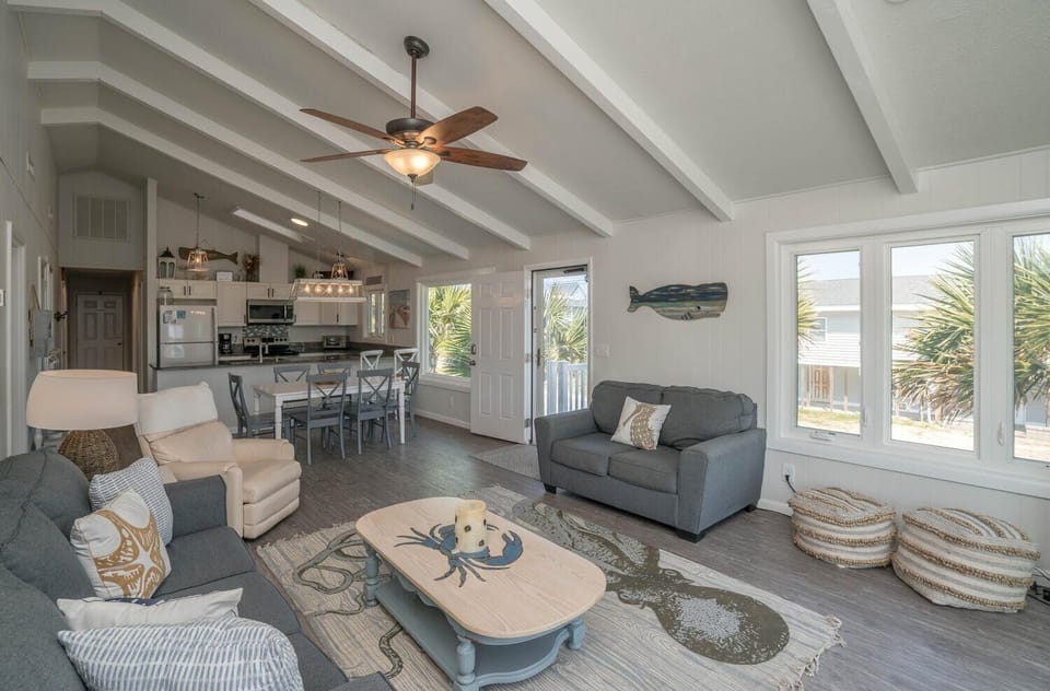 Spacious Oak Island, NC living area with vaulted beam ceilings, ceiling fan, gray sofas, recliner, coffee table, coastal rug, dining table, white kitchen, windows, beach décor, and door opening to a sunny deck.