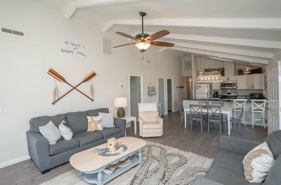 Spacious Oak Island, NC living area with vaulted beam ceilings, ceiling fan, gray sofas, recliner, coffee table, coastal rug, dining table, white kitchen, windows, beach décor, and door opening to a sunny deck.