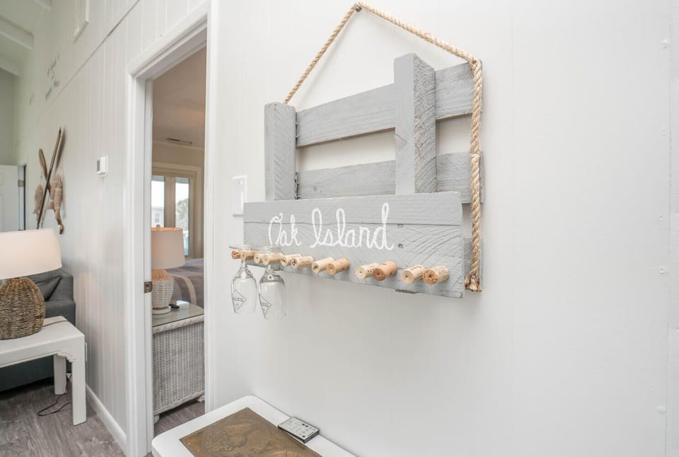 Beachy wall accent featuring a handcrafted Oak Island sign with cork hooks and hanging stemware. Soft coastal tones and rope detailing add character and a relaxed seaside vibe to this inviting nook.
