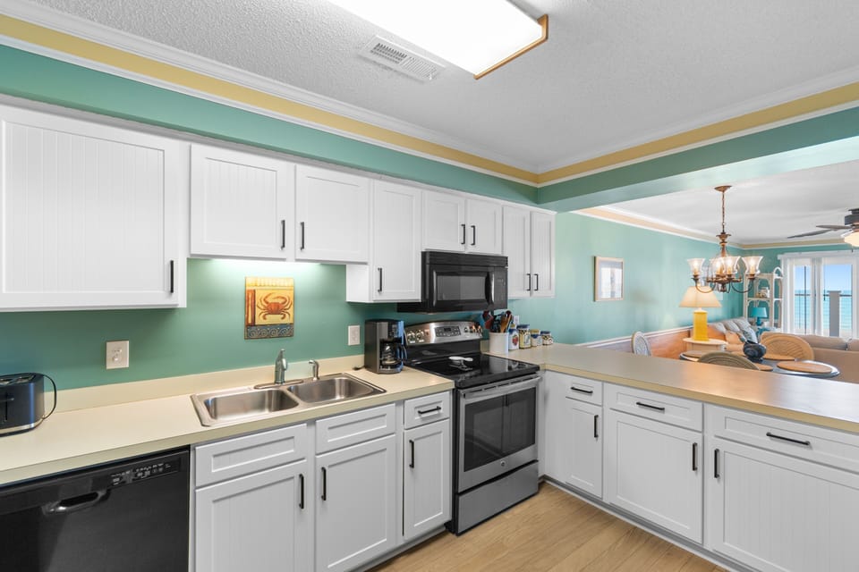 Your fully equipped kitchen with coastal flair welcomes you to prepare meals in comfort, featuring bright white cabinetry, modern appliances, and a cheerful turquoise accent wall that catches stunning ocean views beyond.