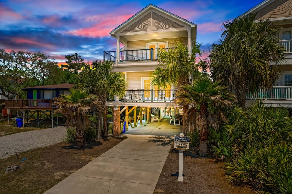 Coastal vacation home with tropical palms and covered parking beneath, framed by spectacular sunset skies.