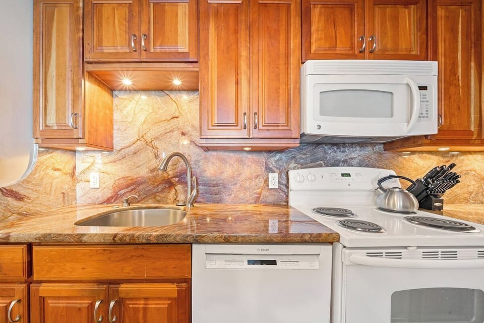 Kitchen with wooden cabinetry, white microwave and stove, dishwasher, and stone backsplash and countertop, a bright and functional space for cooking.
