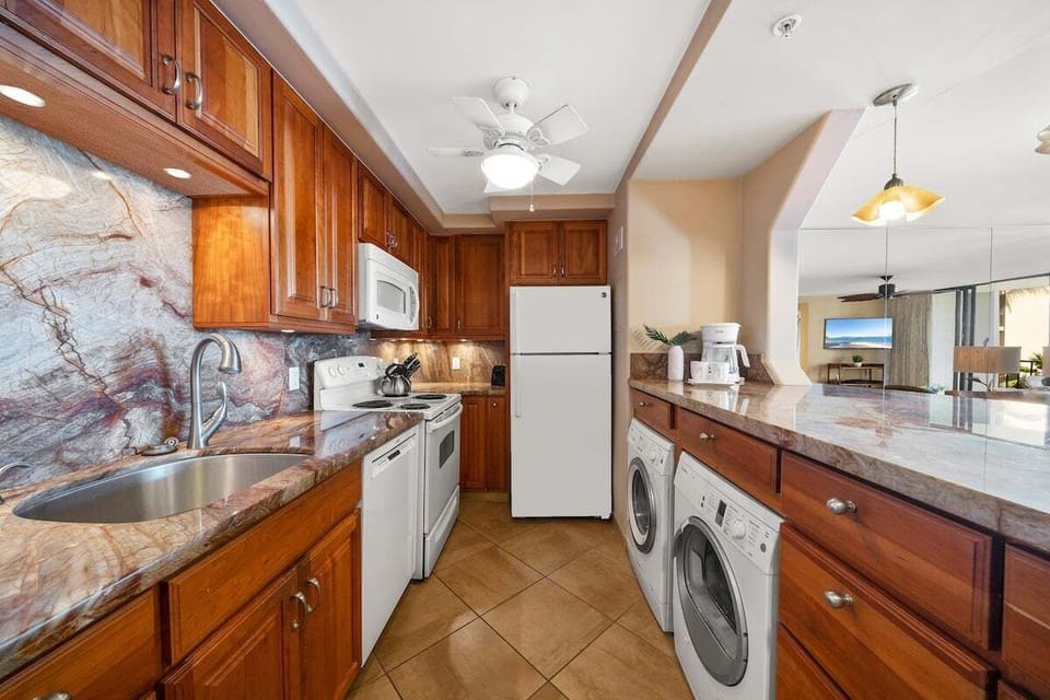 Kitchen with wooden cabinets, granite countertops, and modern appliances including refrigerator, microwave, stove, and washing machine, all warmly lit.