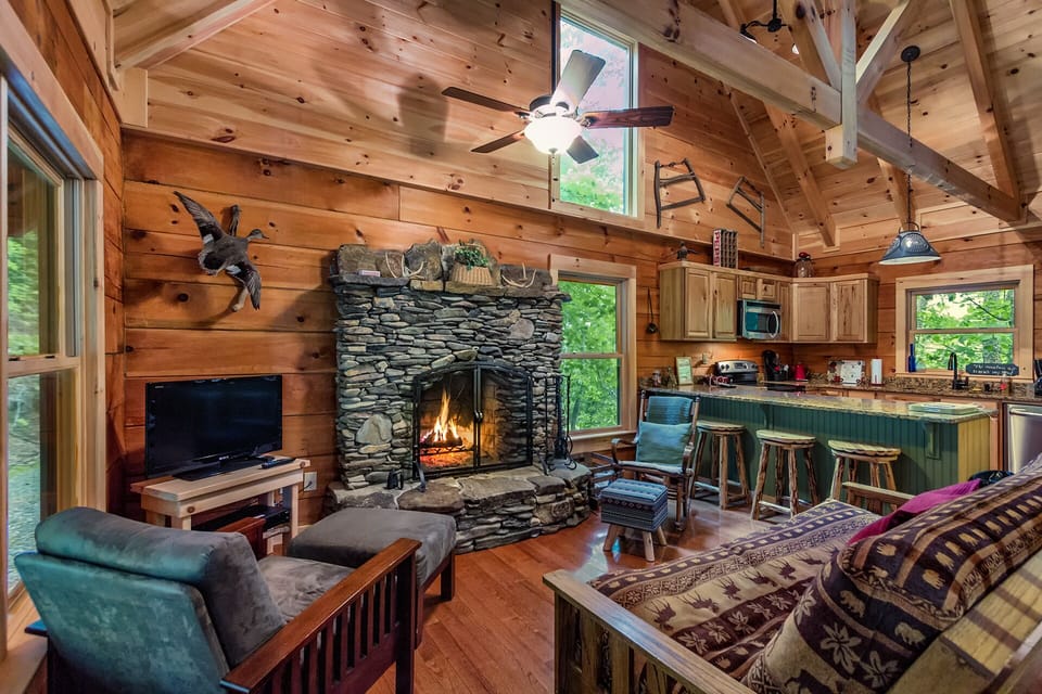 The living room has a modern open floorplan with vaulted beam ceiling, traditional wood walls, a large hand built stacked stone wood burning fireplace and rustic décor throughout. 