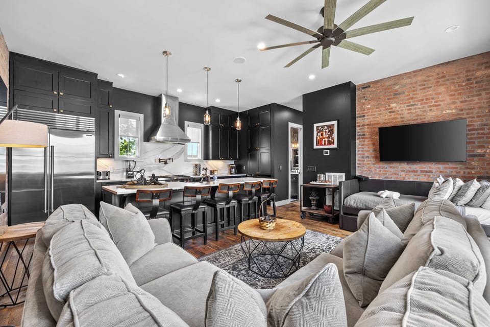 Spacious and inviting living room featuring comfortable sectional seating, exposed brick walls, a Smart TV, and an open-concept layout that flows seamlessly into the kitchen and dining area.
