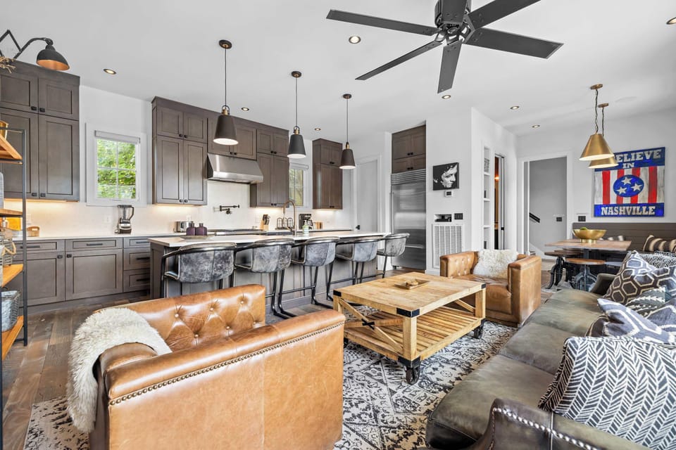 Stylish open-concept living room featuring exposed brick walls, comfortable leather seating, a large Smart TV, rustic wood accents, and a seamless flow into the fully equipped kitchen and dining area.