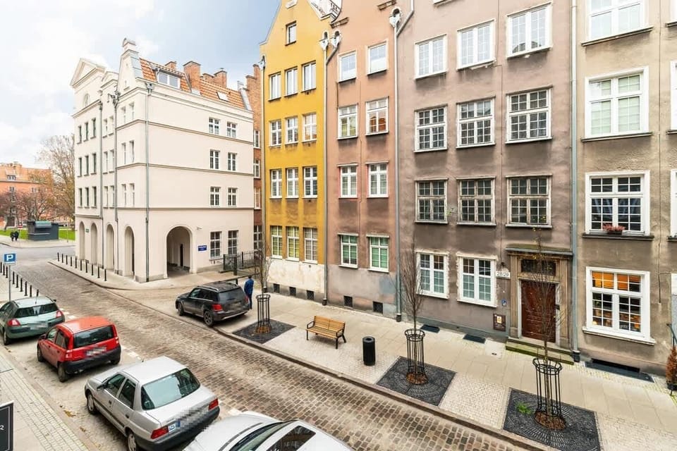 A charming view of a narrow street lined with colorful, historic buildings, typical of European architecture, with cars parked along the cobblestone road.