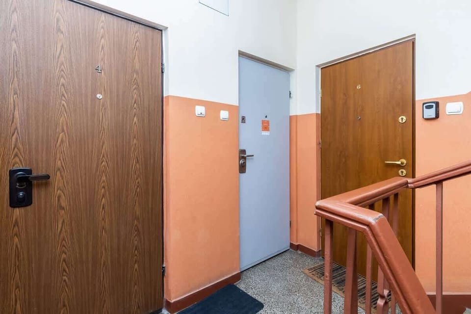  A view of an apartment entrance with two brown doors, one of which leads to a hallway with a staircase, all designed in a simple, clean, and functional style.