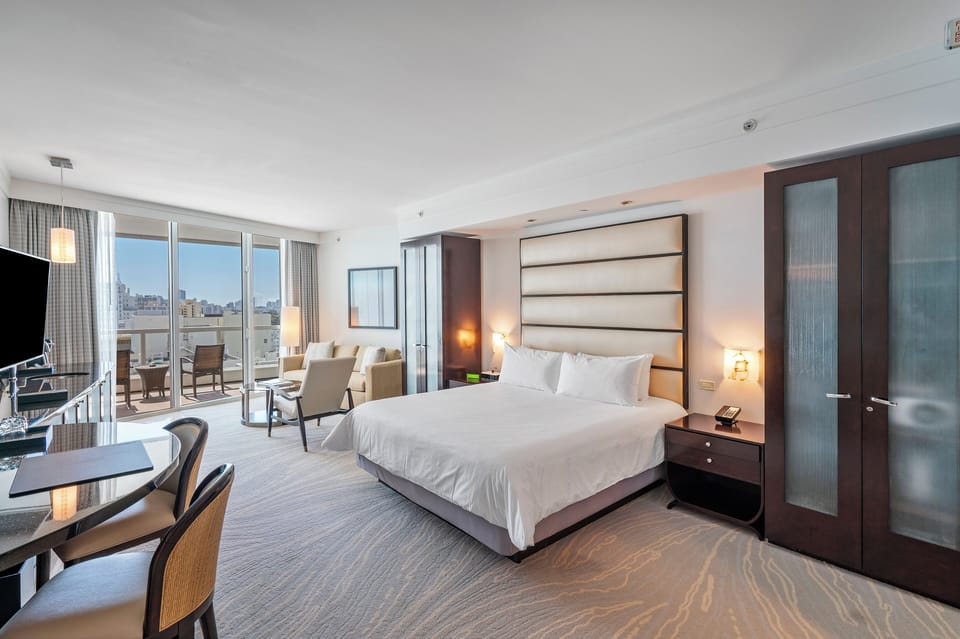 A modern room with a large bed, work desk, seating area, and a balcony. The room features neutral tones and contemporary furnishings with city and ocean views outside the balcony.