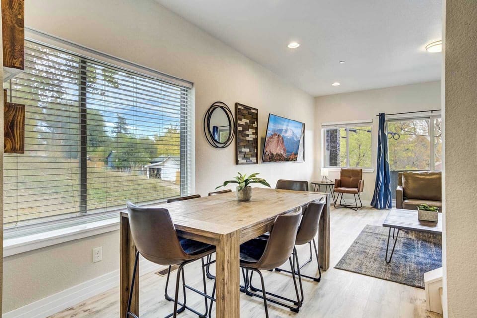 Sunlit dining area with a long table, comfortable chairs, and large windows overlooking greenery. Share unhurried meals or morning coffee here, with natural light and an open layout that invites lingering conversations.