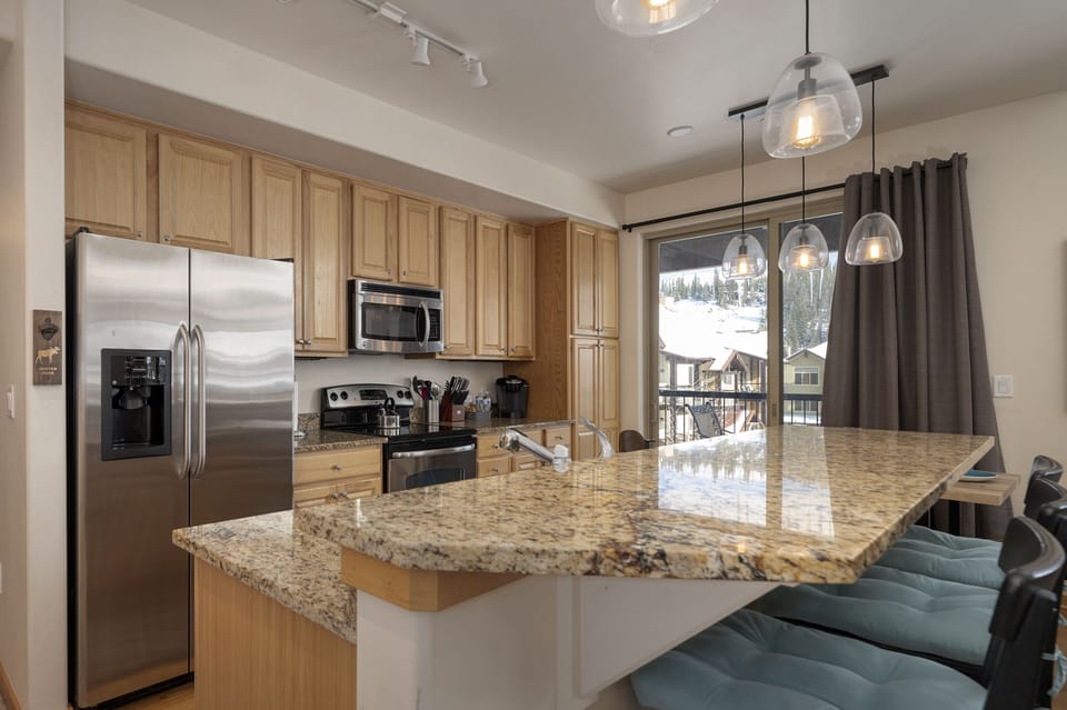 Modern kitchen with wooden cabinets, stainless steel appliances, granite countertop island with teal cushioned bar stools, and pendant lights. Snow-covered landscape visible through large window.