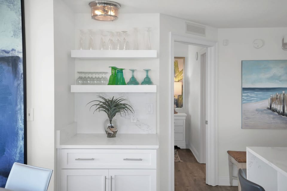 Your bright kitchen features elegant built-in shelving displaying colorful glassware and a charming plant, creating the perfect coastal retreat vibe.