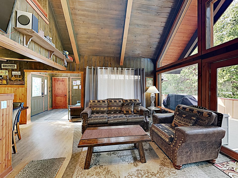 Living room with patterned brown sofa and chair, wood-paneled walls, vaulted ceiling, and large windows overlooking the deck.