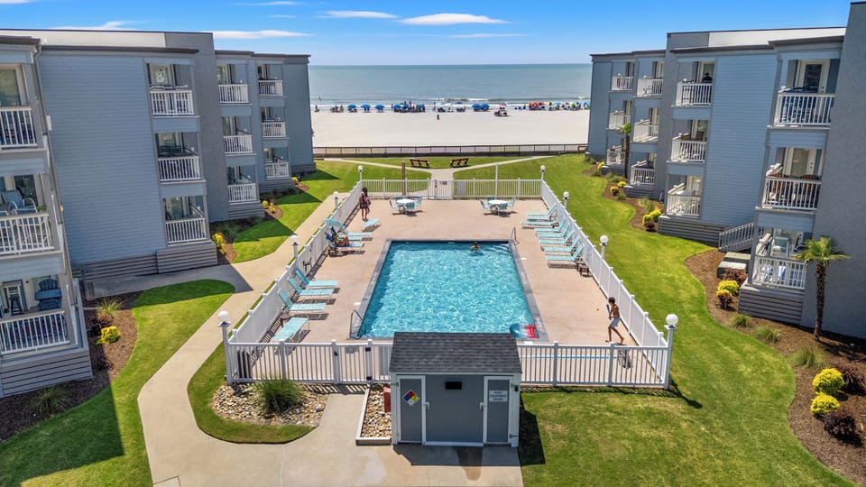 Beachfront property featuring a swimming pool, manicured lawns, and direct access to sandy shores with ocean views, framed by multi-story residential buildings with private balconies.