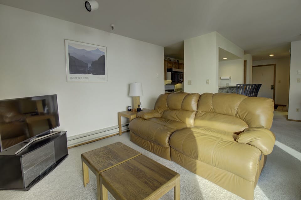A beige living room featuring a leather couch, a flat-screen TV on a stand, a wooden coffee table, and a wall poster. The kitchen and a small dining area are visible in the background.