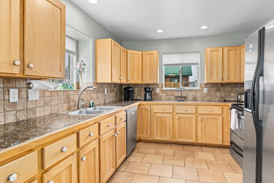 Kitchen with granite countertops, light oak cabinets, tiled backsplash, stainless steel appliances, and two windows.