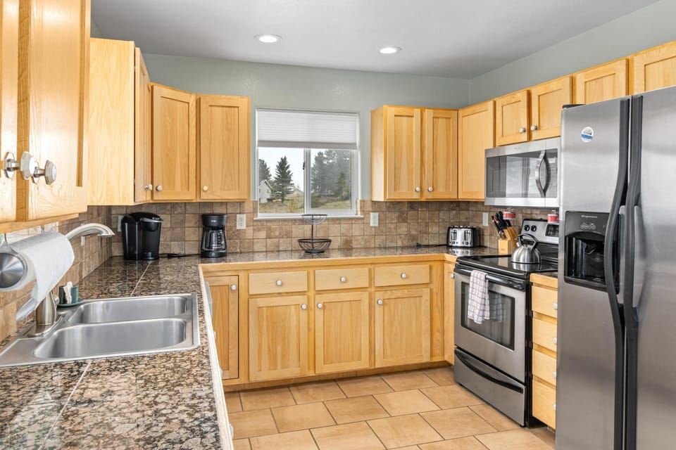 Kitchen with honey oak cabinets, granite tile countertops, stainless steel appliances, and a window overlooking trees.