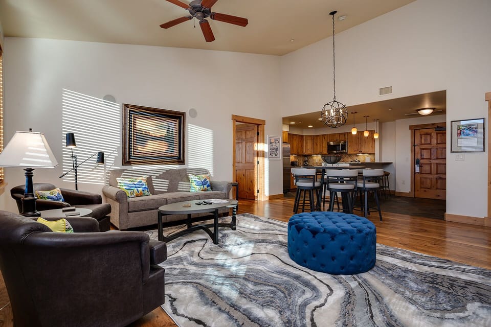 Unwind in your spacious living area where warm sunlight streams through the blinds, highlighting the comfortable seating and open kitchen design.