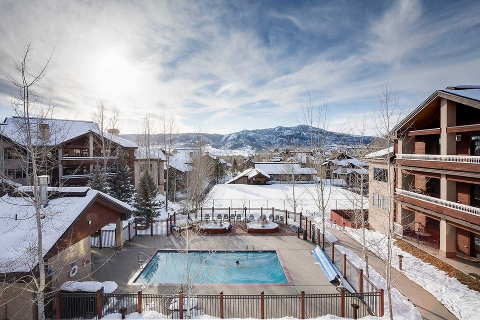 Mountain resort community featuring heated outdoor pool surrounded by snow-covered alpine buildings and scenic mountain backdrop.