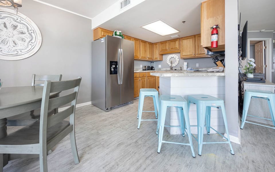 The modern kitchen features warm wood cabinetry, a large stainless refrigerator, and a breakfast bar with blue stools