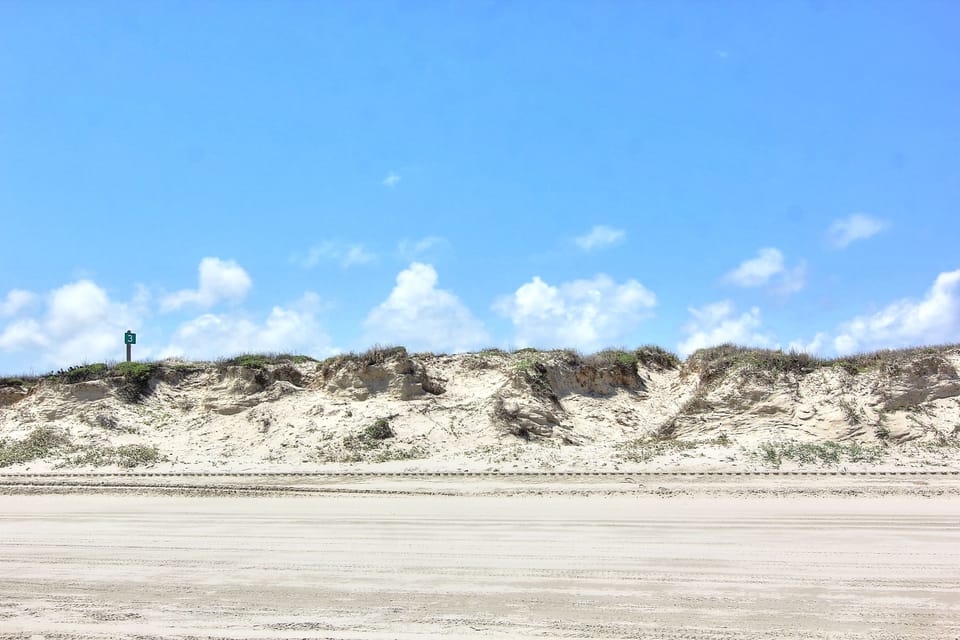 Natural coastal dunes rise above pristine sandy shoreline, creating a peaceful beachfront setting with clear blue skies.